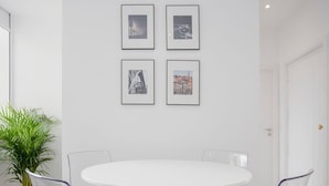 A modern dining area with a white round table and four transparent chairs with metal frames. There are four framed photographs on the wall above the table. - Liiiving in Porto | Center Spot Apartment (Porto)