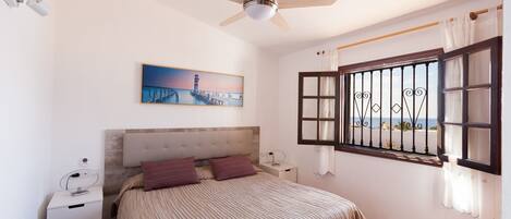 A bedroom with a bed, a nightstand, a ceiling fan, a window with bars, and a painting on the wall.