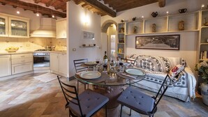 A dining area with a glass table, chairs, and a sofa. There is a kitchen in the background with cabinets and a stove. - La Casina di Pia in Alta Maremma (Montieri)