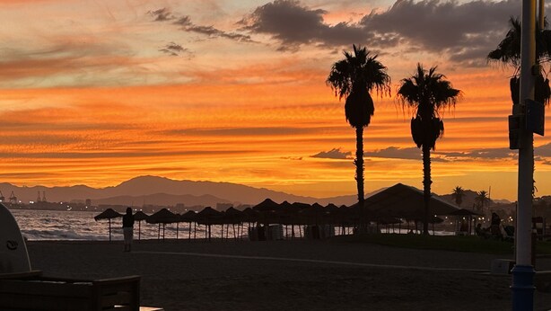 Een strandscène met palmbomen, strandparasols en een zonsondergang.