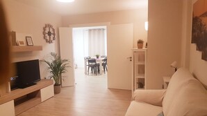 Een woonkamer met een televisie, een plant en een eethoek met een tafel en stoelen.