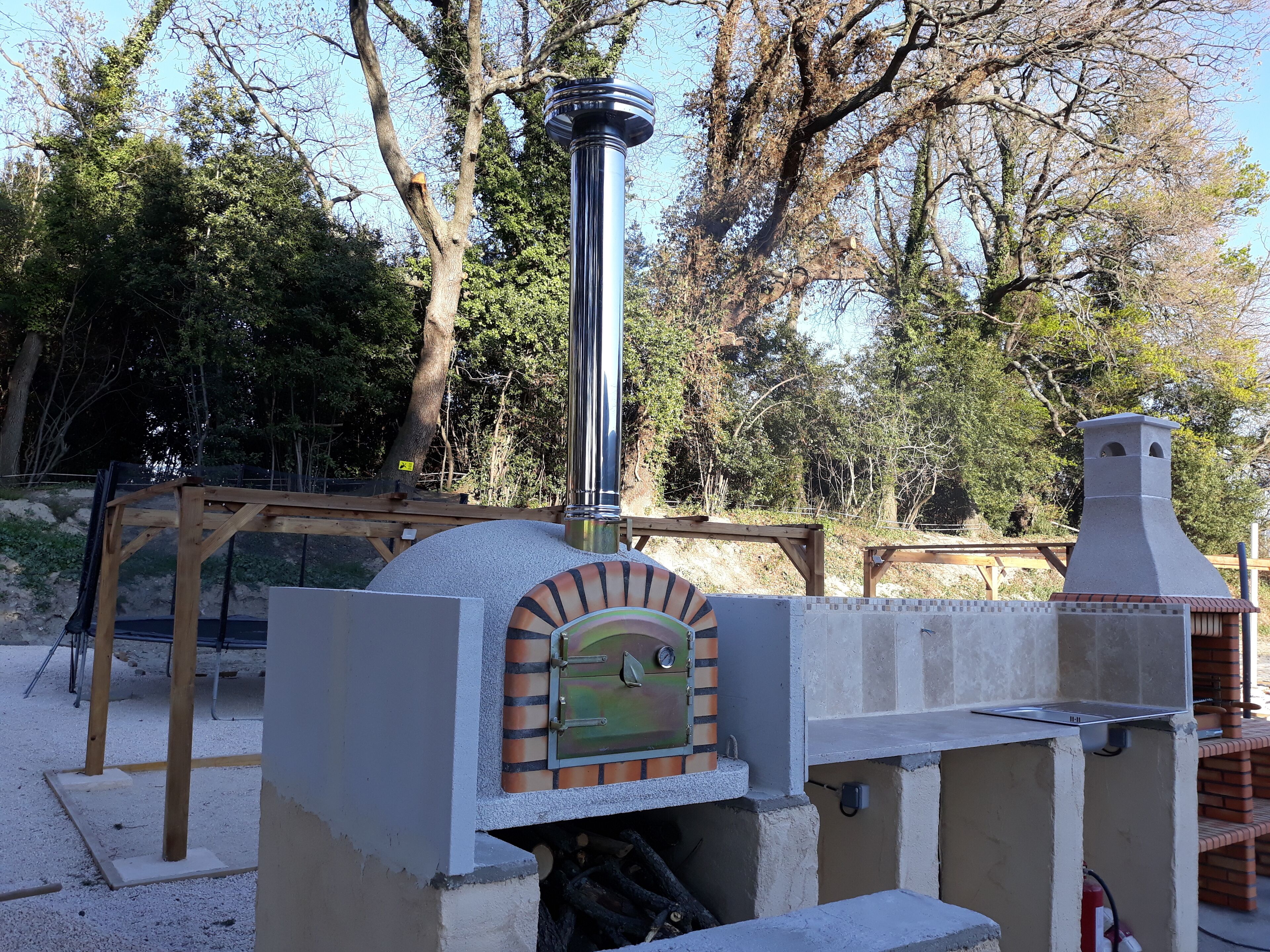 A brick oven with a metal chimney, surrounded by trees and a wooden structure.