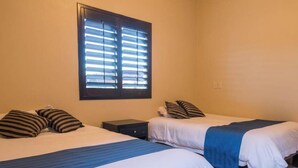 A room with two beds, each with striped pillows and a blue blanket. There is a window with blinds, a nightstand, and a wall socket. - Beautiful house in San Carlos (SAN CARLOS)