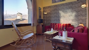 A room with a rocking chair, a red sofa with striped cushions, a small table with a candle and glass, and a stone wall.