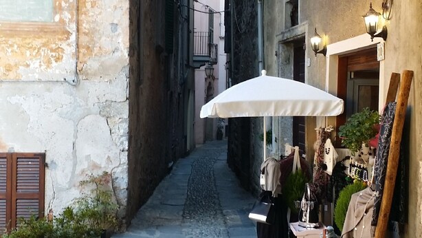 A narrow cobblestone alleyway with an archway, a white umbrella, and a small outdoor seating area with a table and chairs.