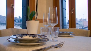 Une table à manger dressée avec des assiettes, des couverts et des verres, agrémentée d’une plante en pot et offrant une vue sur des bâtiments par une fenêtre.