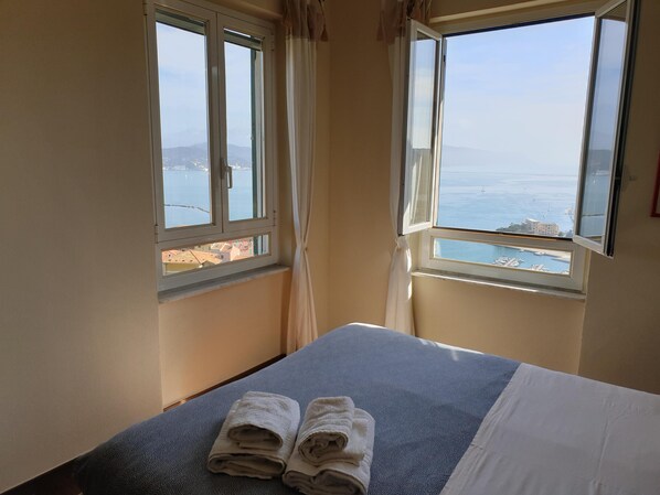 A hotel room with a bed, two towels, and two windows offering a view of the sea and buildings.