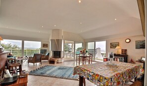 A spacious living area with a large dining table, comfortable seating, a fireplace, and large windows offering a view of the outdoors.