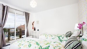 Une chambre avec une grande fenêtre, un lit recouvert d’une couvre-lit à motifs floraux, une table de chevet et une lampe.