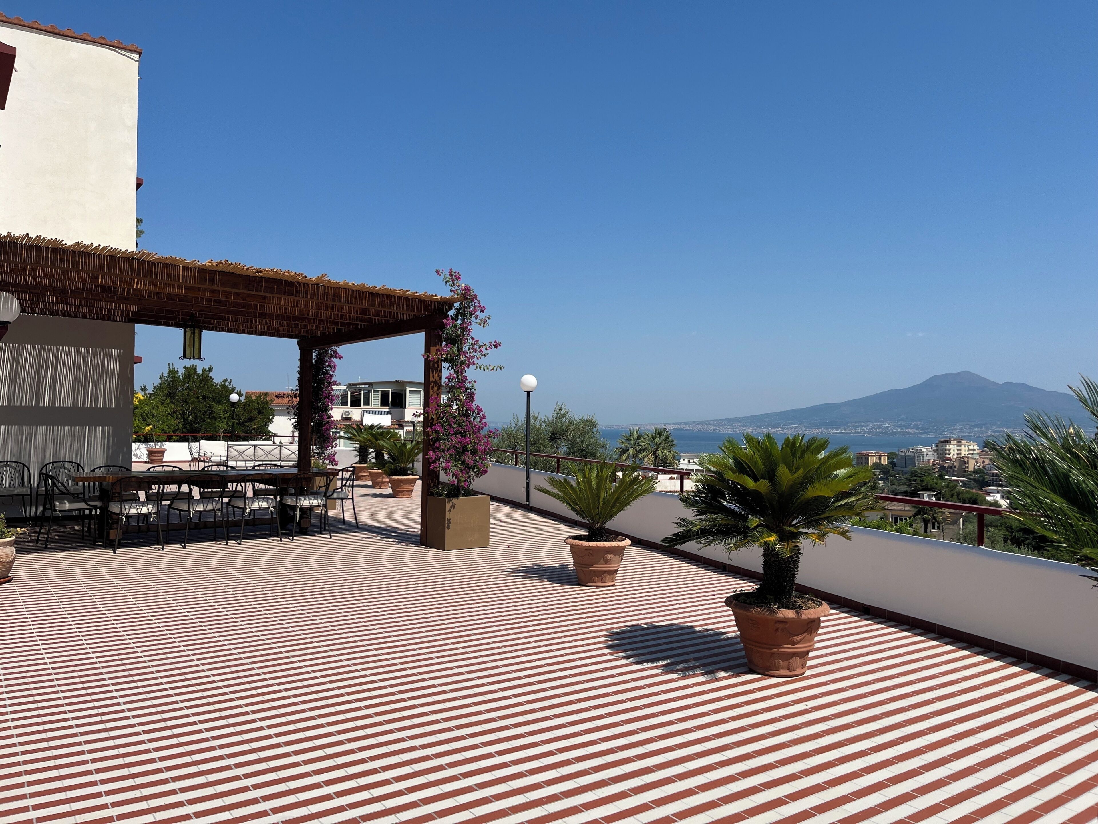 Eine Dachterrasse mit Backsteinpflaster, Außenmöbeln, Blumentöpfen und Blick auf Berge und Stadt.