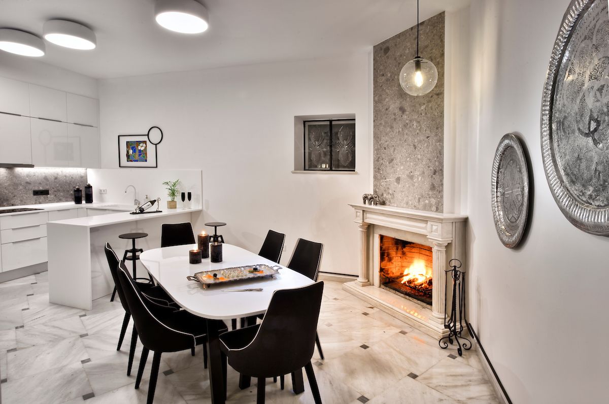 A modern kitchen with a central island, dining table, and chairs. There is a fireplace with a visible fire, and decorative plates on the wall.