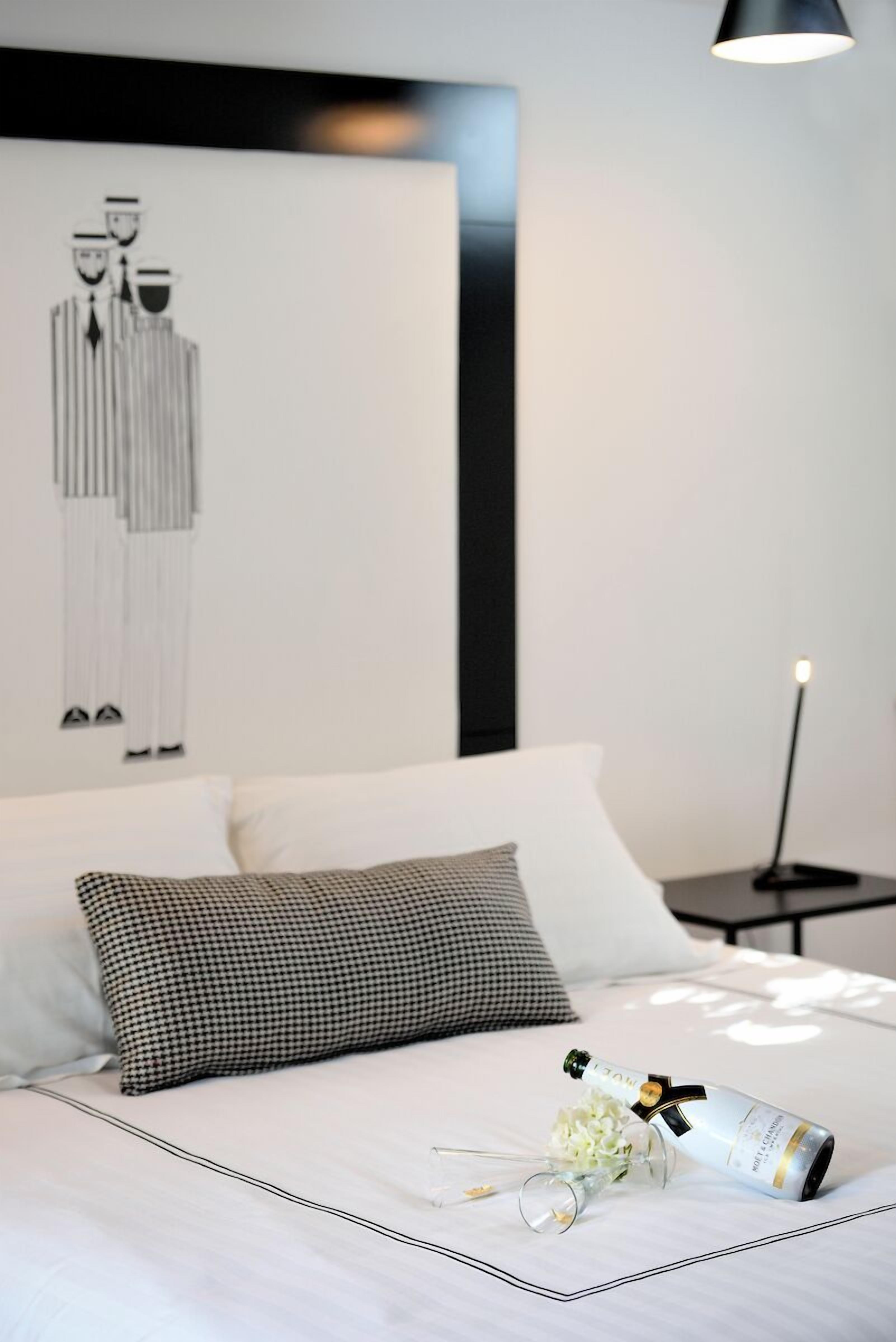 A neatly made bed with a champagne bottle and flowers, a black and white artwork on the wall, and a bedside table with a lamp.