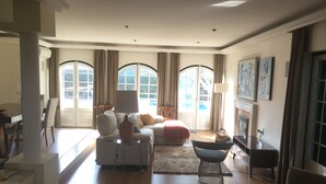 A spacious living room with a sofa, armchair, and a coffee table. There is a fireplace and a dining area visible.
