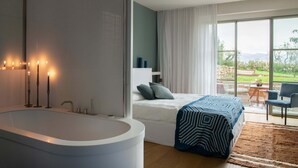 Une chambre moderne avec une grande baignoire, un lit recouvert d’une couvre-lit à motifs et une vue sur un jardin grâce à de grandes fenêtres.