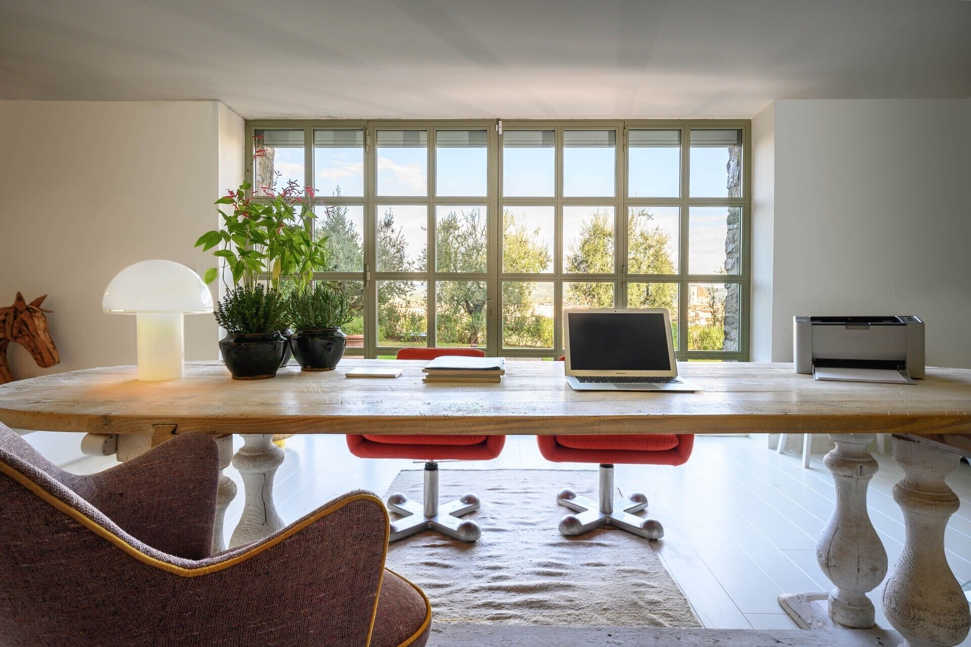 A room with a large window, a laptop, a printer, and two potted plants on a wooden table.