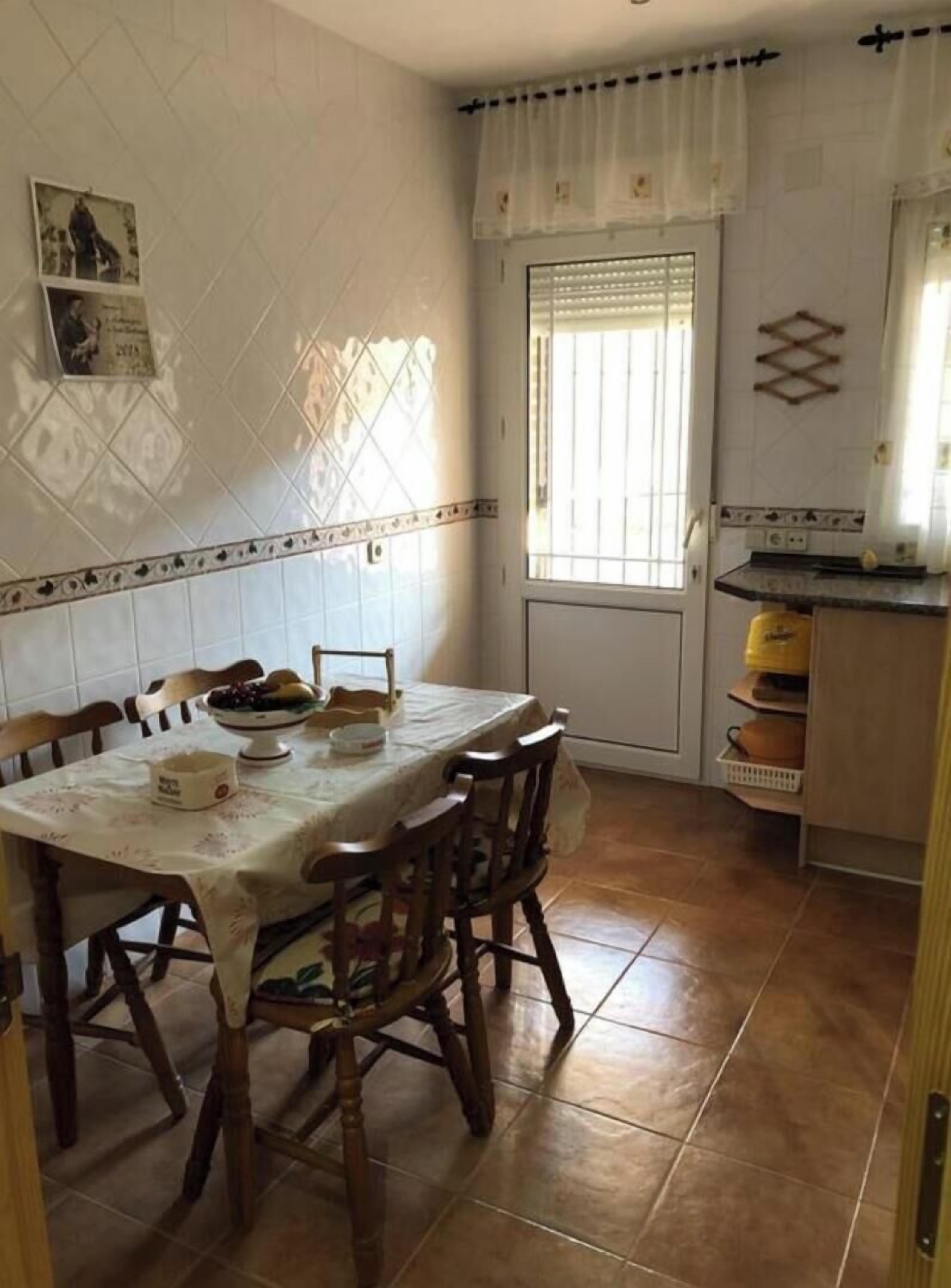 Un coin repas avec une table dressée pour quatre personnes, des chaises en bois, un sol carrelé, une fenêtre avec des rideaux et un espace cuisine équipé d’une cuisinière et de placards.
