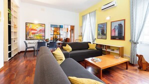 Un salon comprenant un canapé, une table basse et un coin repas avec des chaises et une table. On y trouve également une bibliothèque et un tableau accroché au mur.