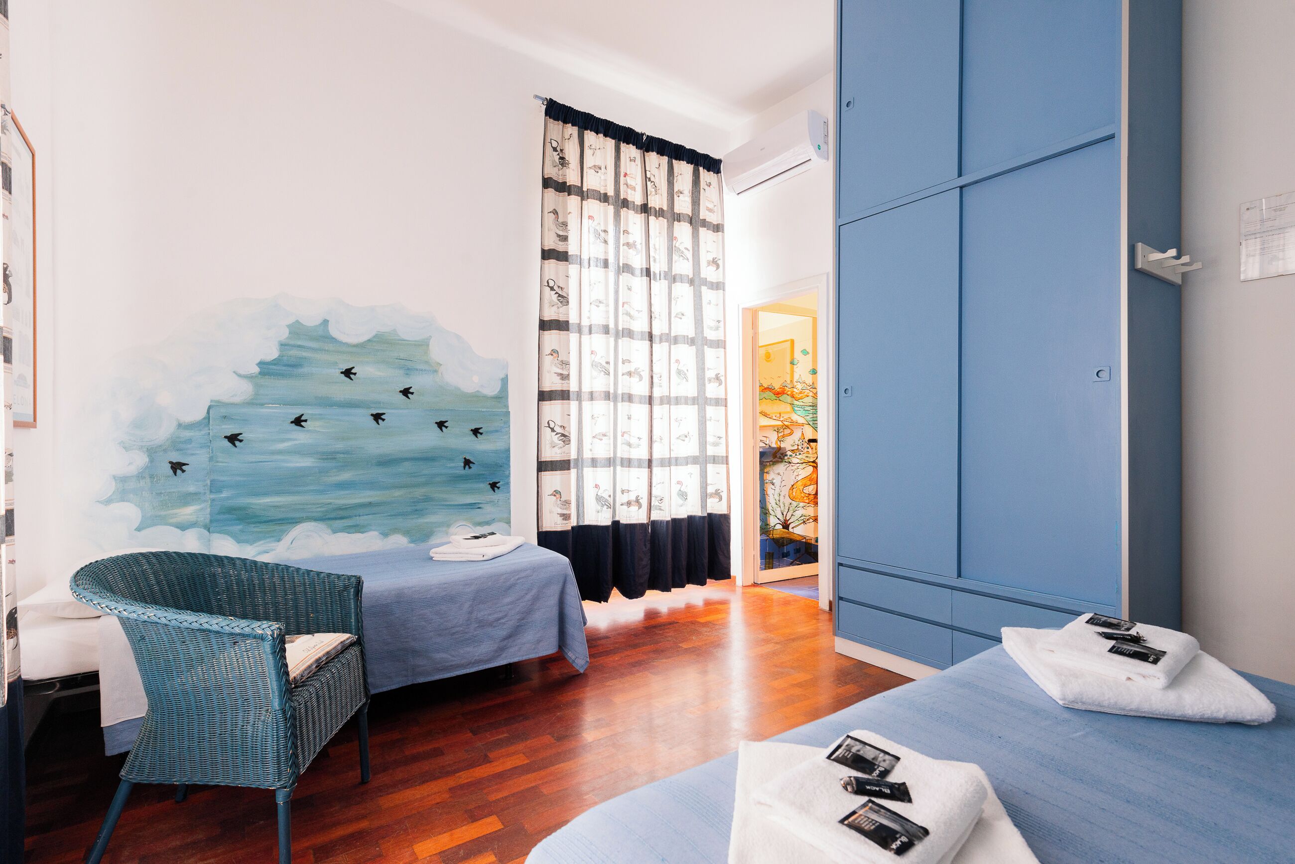 Quarto com cama, uma cadeira e mesa de massagem. Há um mural na parede e uma janela com cortinas.