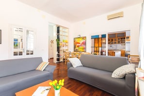 A living room with two grey sofas, a coffee table with a magazine and a vase of yellow tulips, and a kitchen visible through an open door.