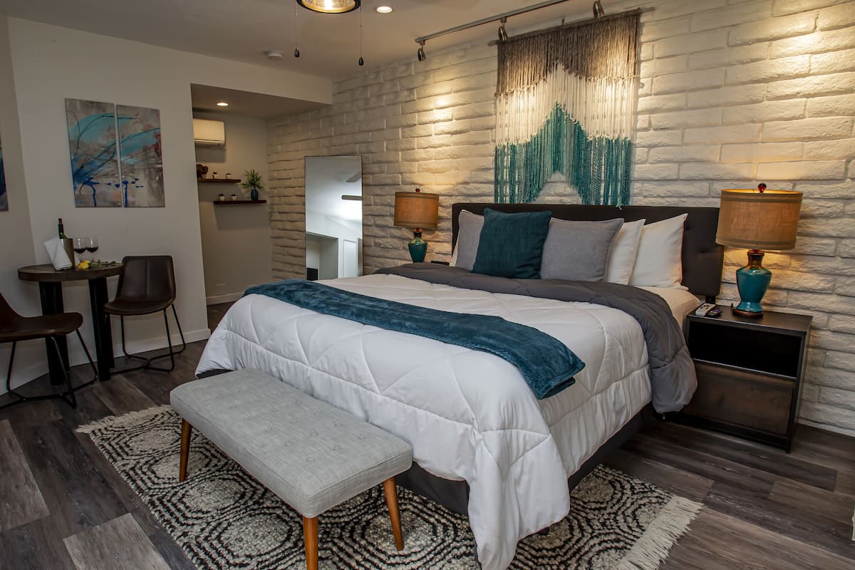 A modern bedroom with a large bed, two bedside tables with lamps, a bench, a desk with chairs, and a wall-mounted artwork.