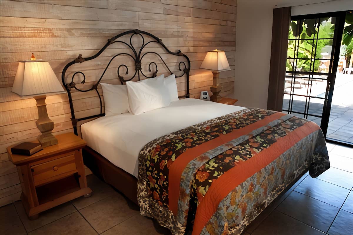 A neatly made bed with a floral bedspread, two bedside lamps, a wooden nightstand, and a view through a glass door to an outdoor area.