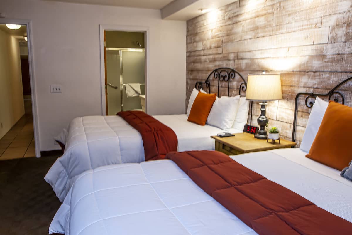 A hotel room with two beds, a wooden accent wall, a nightstand with a lamp, and a visible bathroom door.