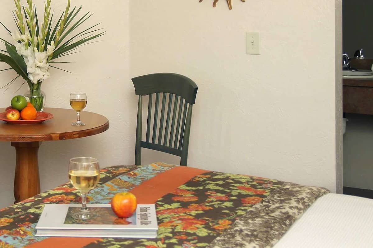 A dining area with a table set for two, featuring a floral tablecloth, a vase with flowers, and a plate with fruits and a glass of wine.