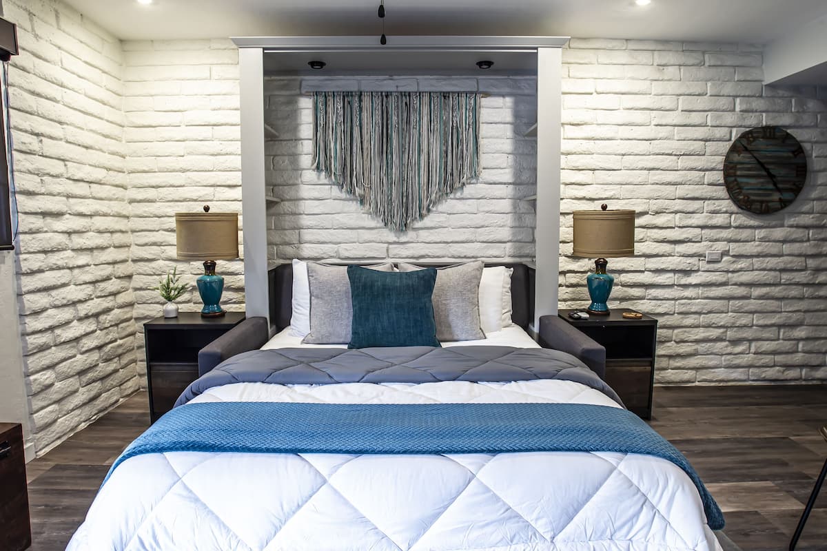 A bedroom with a large bed, white brick walls, a hanging tapestry, two bedside tables with lamps, and a round clock on the wall.