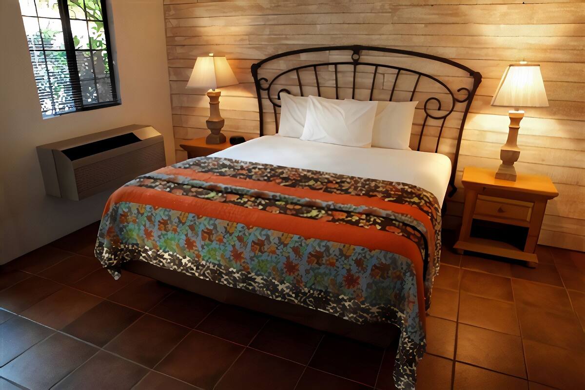 A neatly made bed with a patterned bedspread, two bedside lamps, and a window with blinds.