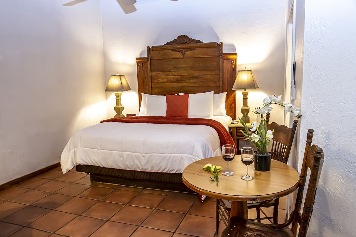 A bedroom with a bed, two bedside lamps, a small round table with a wine glass and flowers, and a chair.