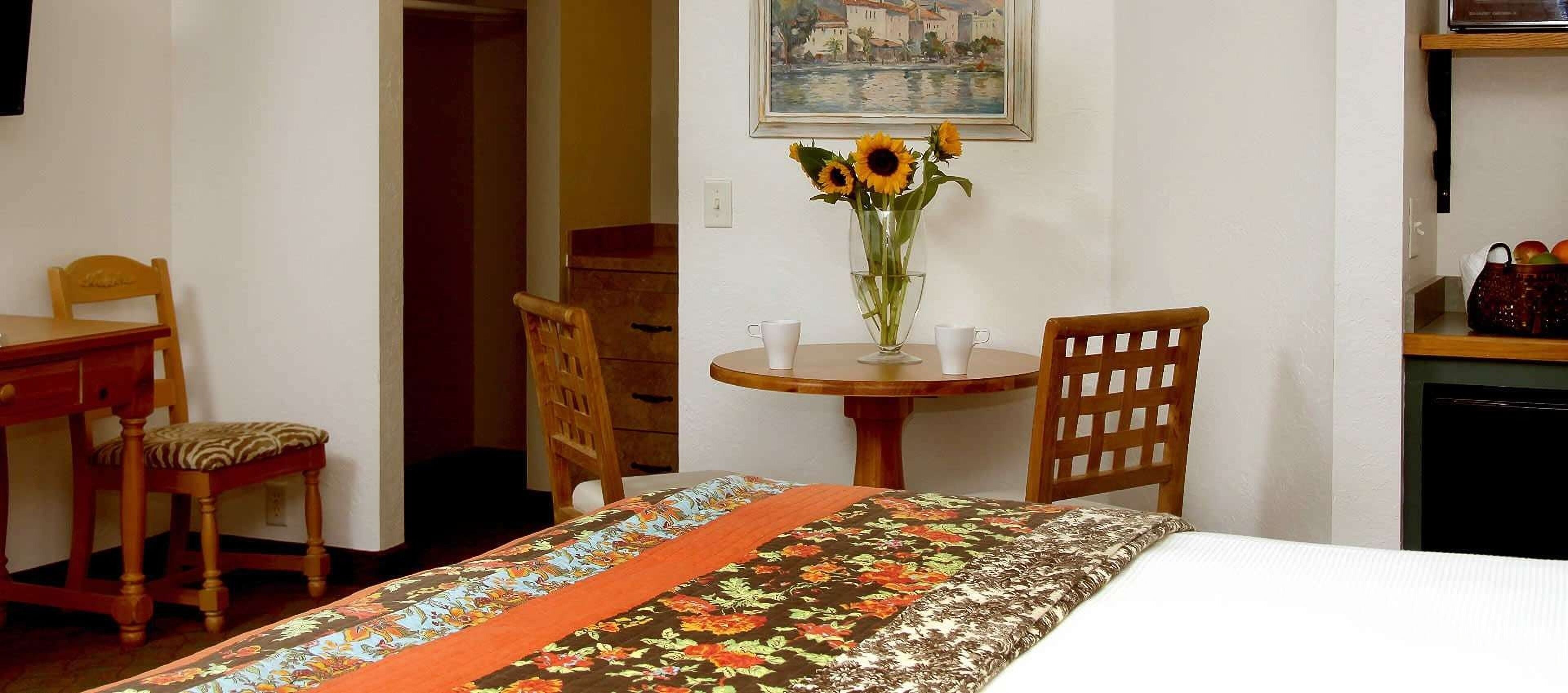 Ein Zimmer mit einem Bett, einem Holzhocker, einem Tisch mit einer Vase voller Sonnenblumen und einem Gemälde an der Wand.
