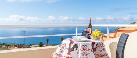 A table with a decorative tablecloth, a bottle, and a bowl of fruit on a balcony overlooking a coastal view.