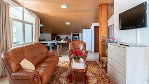 A living room with a leather sofa, a wooden coffee table with a potted plant, a TV on the wall, and a kitchen in the background. - Apartment Lucy , Sunning views over sea and mountains (Arco Da Calheta)