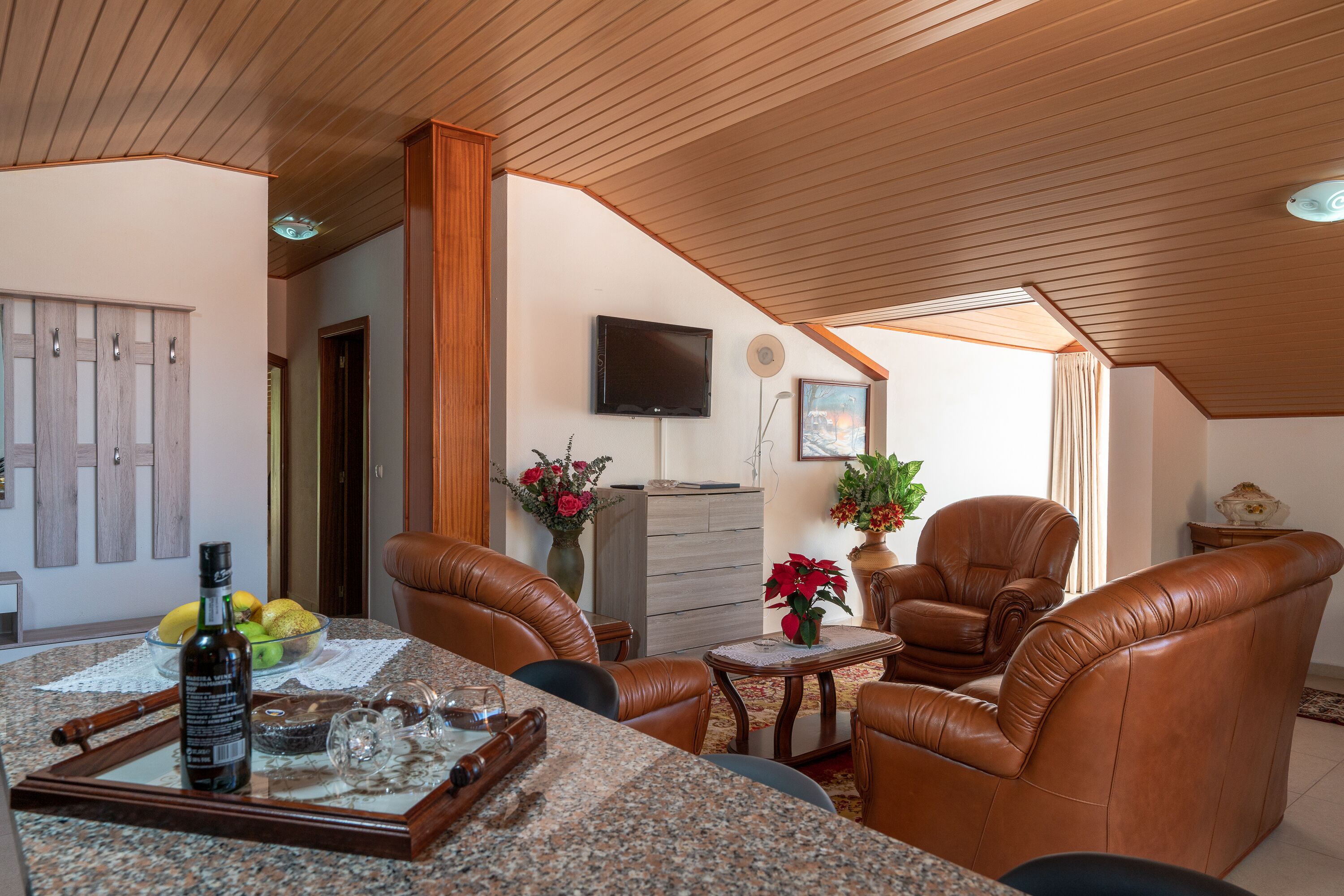 A cozy living room with leather chairs, a dining table with a bottle, glasses, and a bowl of fruit. There's a TV mounted on the wall and a potted plant on a small table.