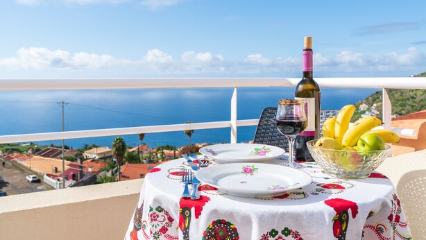 A table set with plates, a wine bottle, and a bowl of fruit on a balcony overlooking the sea. - Apartment Lucy , Sunning views over sea and mountains (Arco Da Calheta)