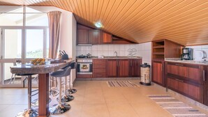 A modern kitchen with wooden cabinets, a central island with bar stools, a coffee machine, and a view through large windows. - Apartment Lucy , Sunning views over sea and mountains (Arco Da Calheta)