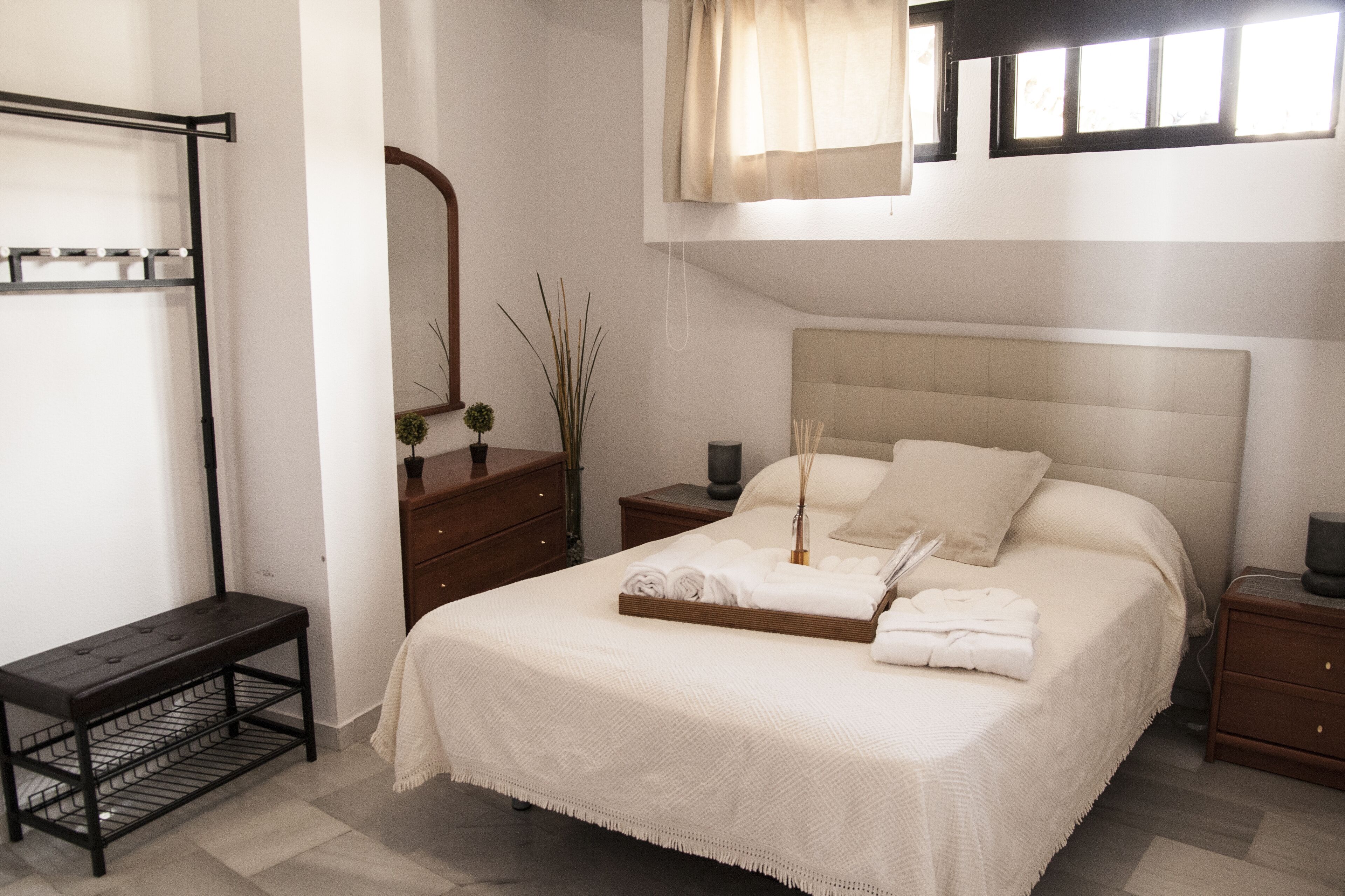 A bedroom with a bed, bedside tables, a bench, a mirror, and a window with curtains.
