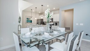 A modern dining area with a glass table set for six, white chairs, and a vase with white flowers. - Stylish and Stunning 5BD Pool Home in Solterra - #5st452 (Davenport)