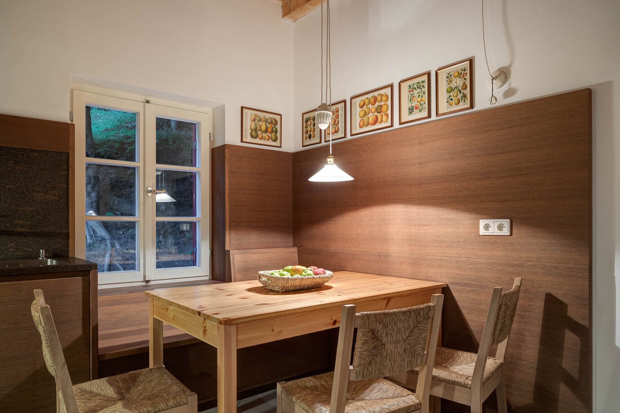 A wooden dining table with chairs, a window with a view of trees, and framed fruit illustrations on the wall.