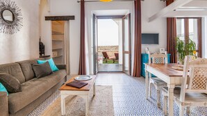 A living room with a sofa, coffee table, and a dining area with a table and chairs. There is a large window with curtains, a ceiling light, and a wall-mounted TV. - Studio gound floor with garden and Sea Views (Tarifa)