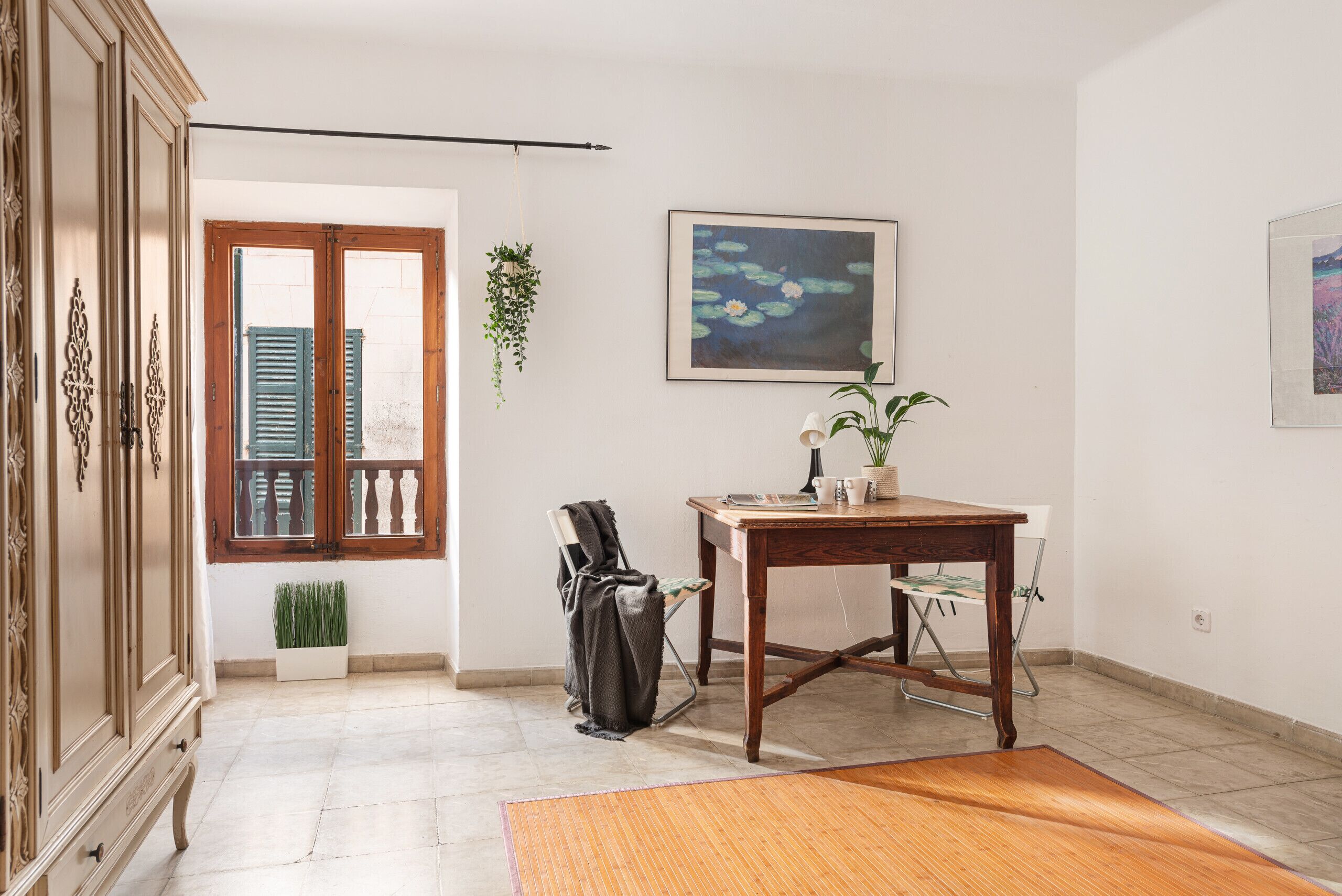 A room with a wooden table, a chair, a radiator, a hanging plant, and a framed picture on the wall.
