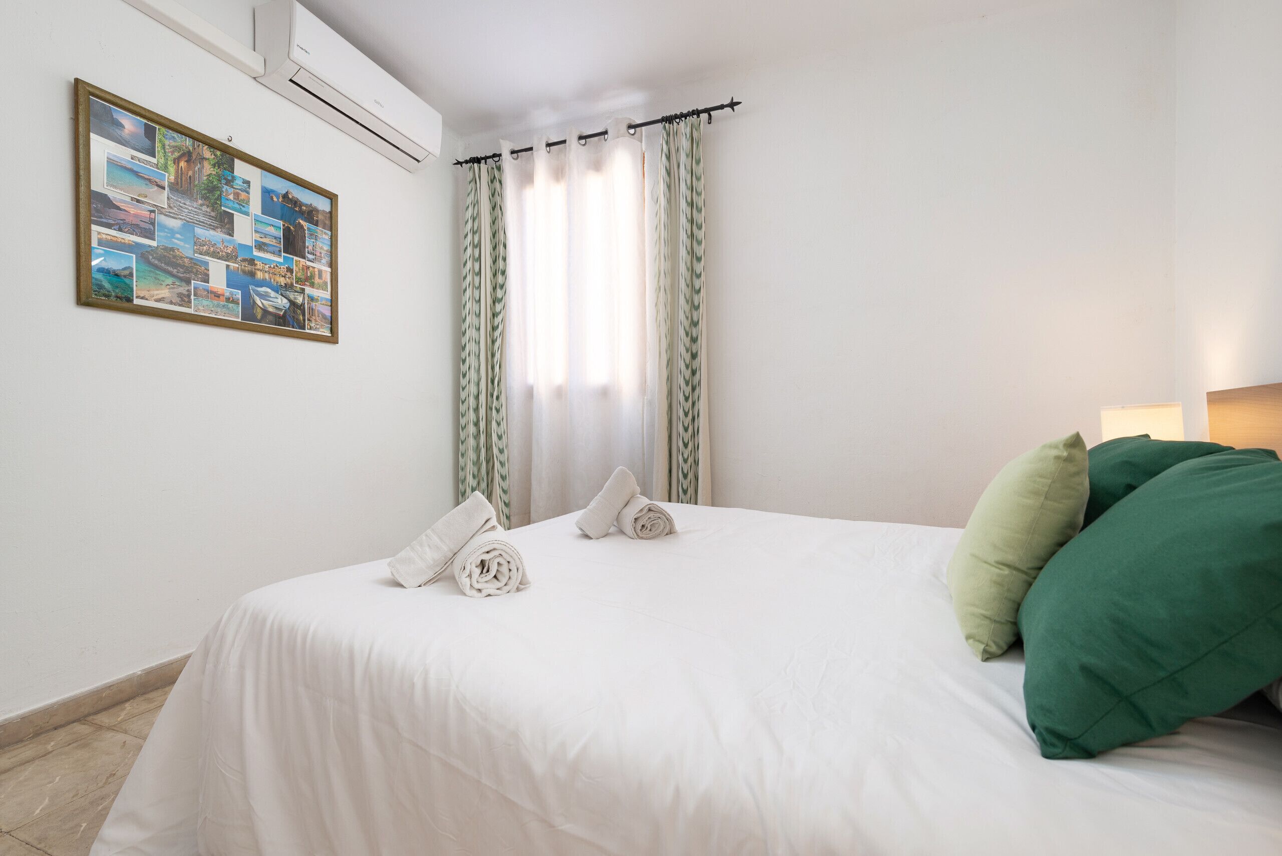 Una cama bien hecha con ropa blanca, dos toallas enrolladas y almohadas verdes. Un collage enmarcado de fotografías está montado en la pared sobre la cama.