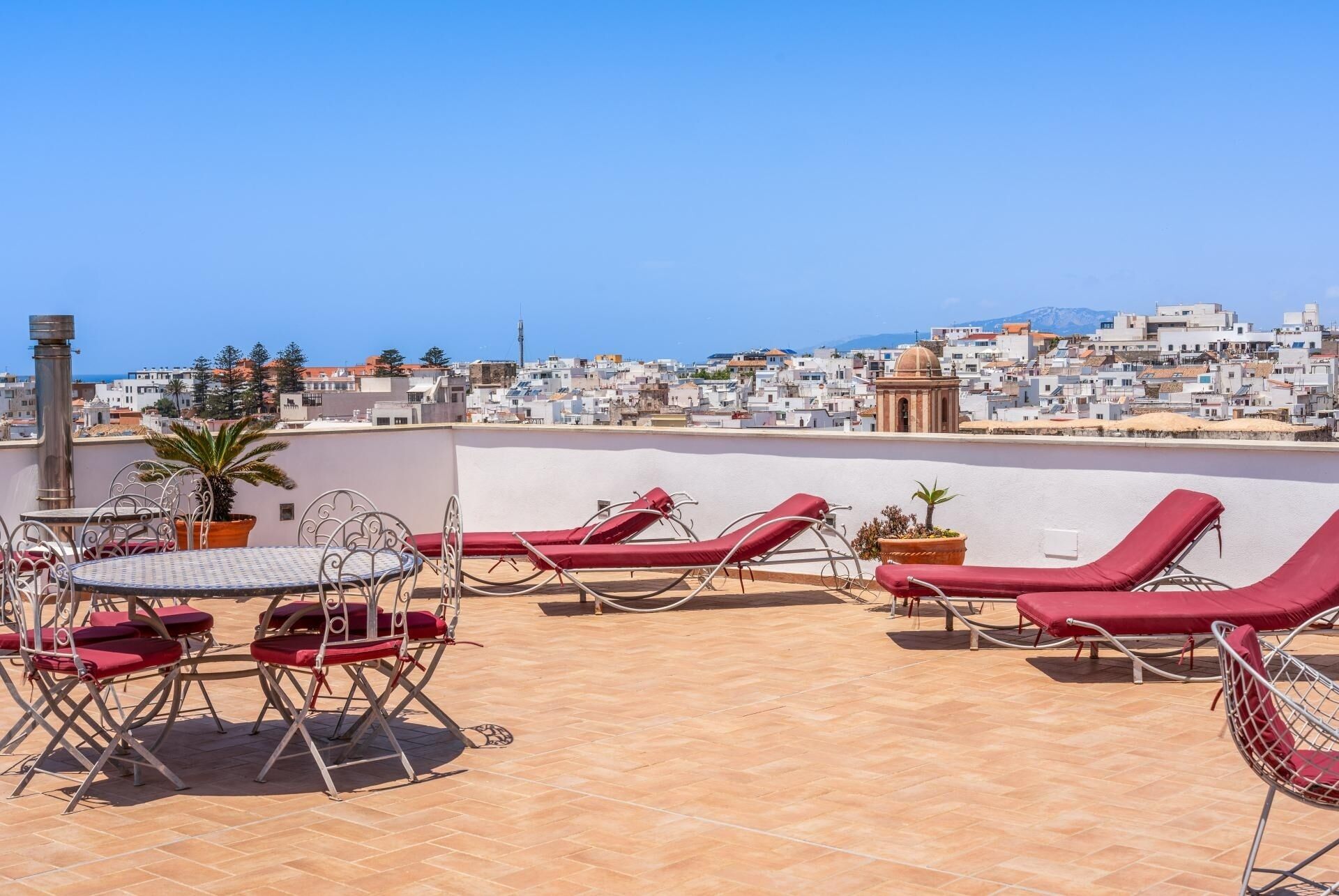 A rooftop terrace with red lounge chairs and a metal table, overlooking a coastal cityscape.