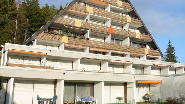 A multi-story building with balconies, a covered outdoor seating area, and a garage entrance. - Unwind, relax, recover ... - Indoor swimming pool (12x6m) u. Sauna in the house! (Unterkirnach)