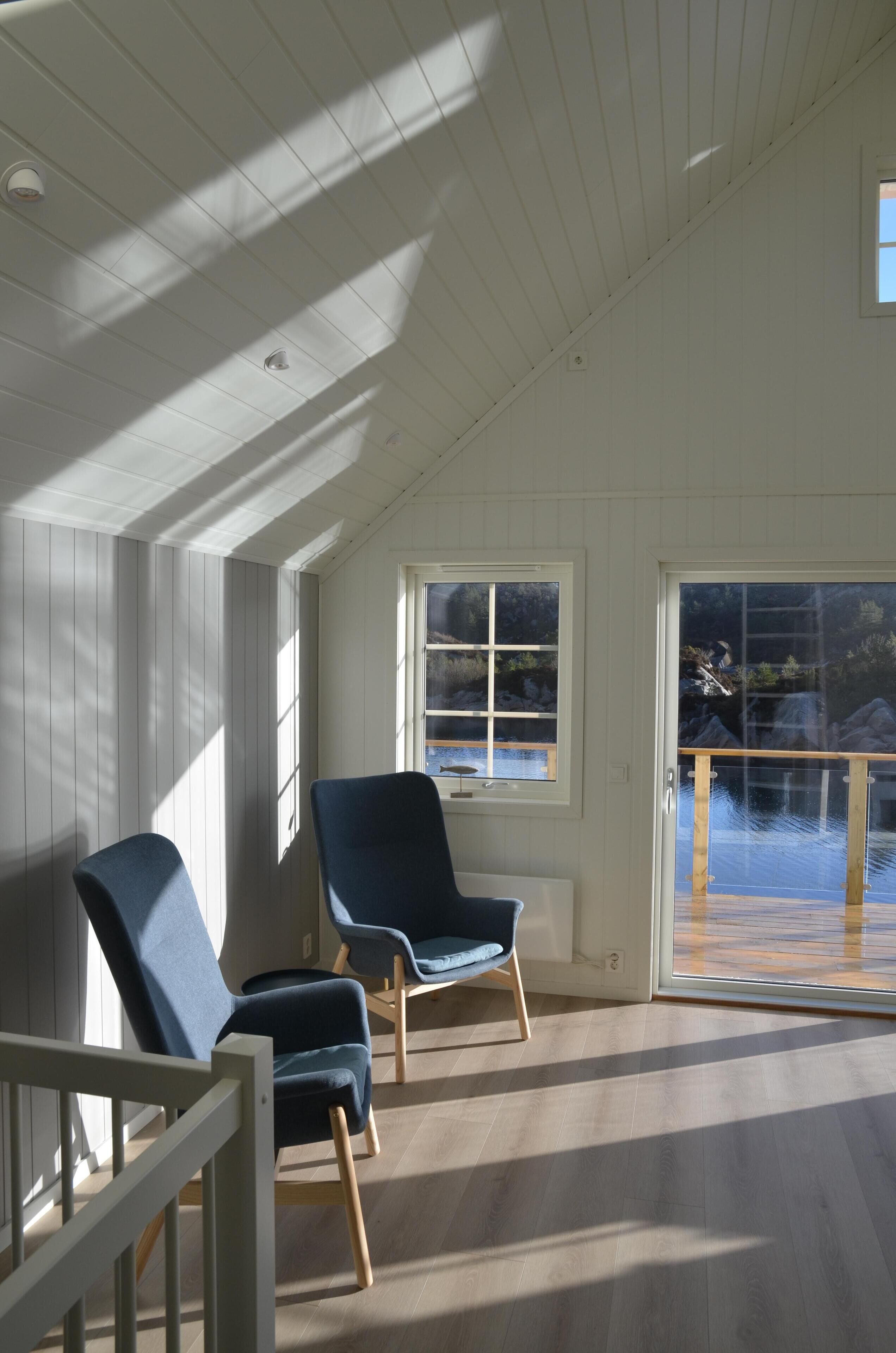 A room with two blue chairs, a wooden floor, and a view of a water body through a glass door.