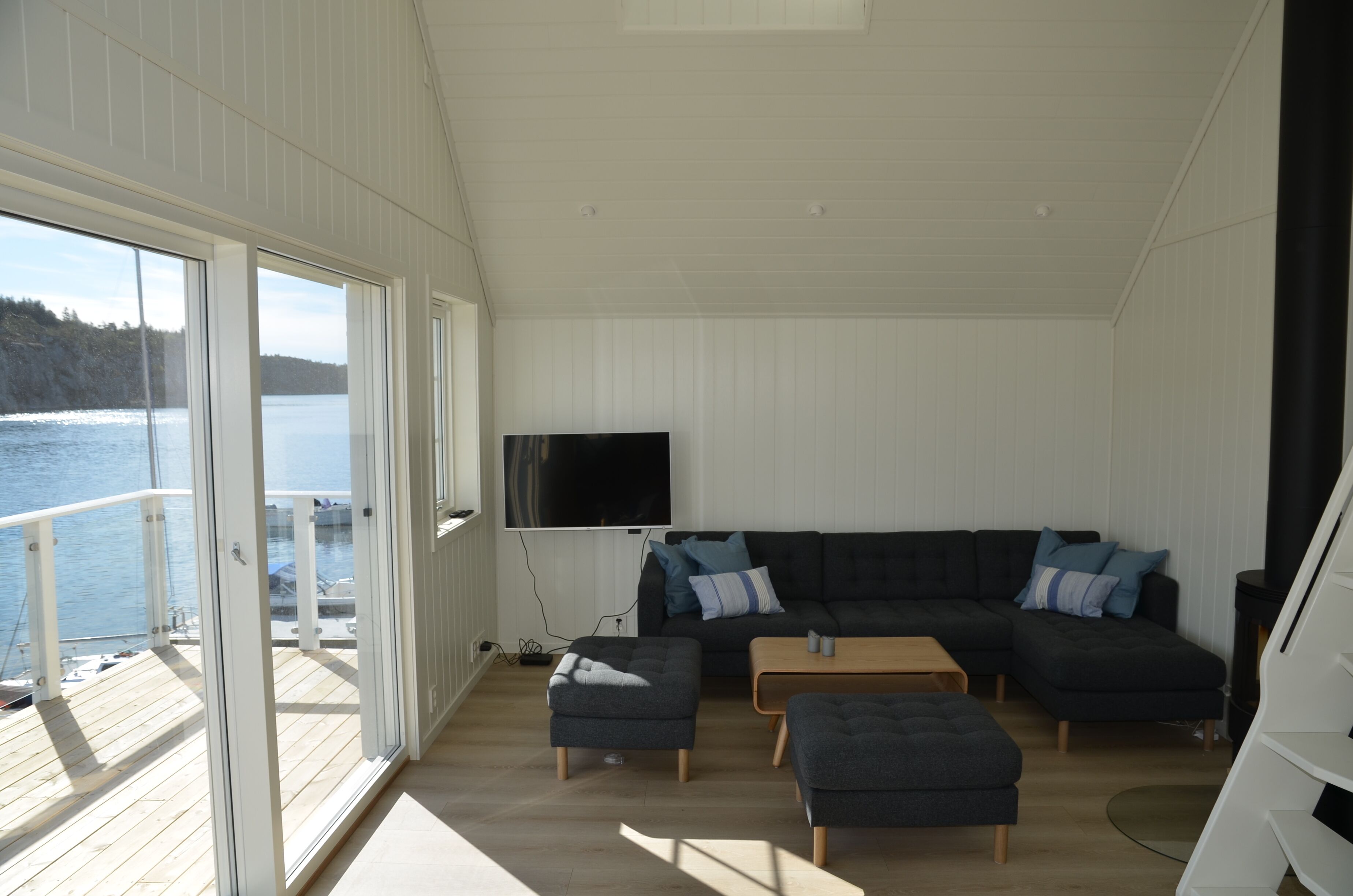 A modern living room with a large sectional sofa, two ottomans, and a coffee table. A flat-screen TV is mounted on the wall. The room has large windows offering a view of a lake and forested hills.
