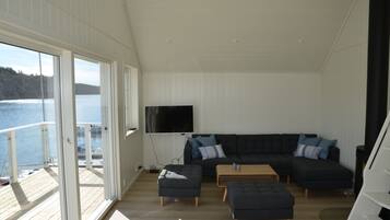 A modern living room with a large sectional sofa, two ottomans, and a coffee table. A flat-screen TV is mounted on the wall. The room has large windows offering a view of a lake and forested hills.