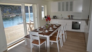 A dining area with a wooden table set for six, white chairs, and a view of a lake through large glass doors. - Fishing, 2 top boats, swimming, SUP, workation, relaxing, everything is possible (Urangsvåg)