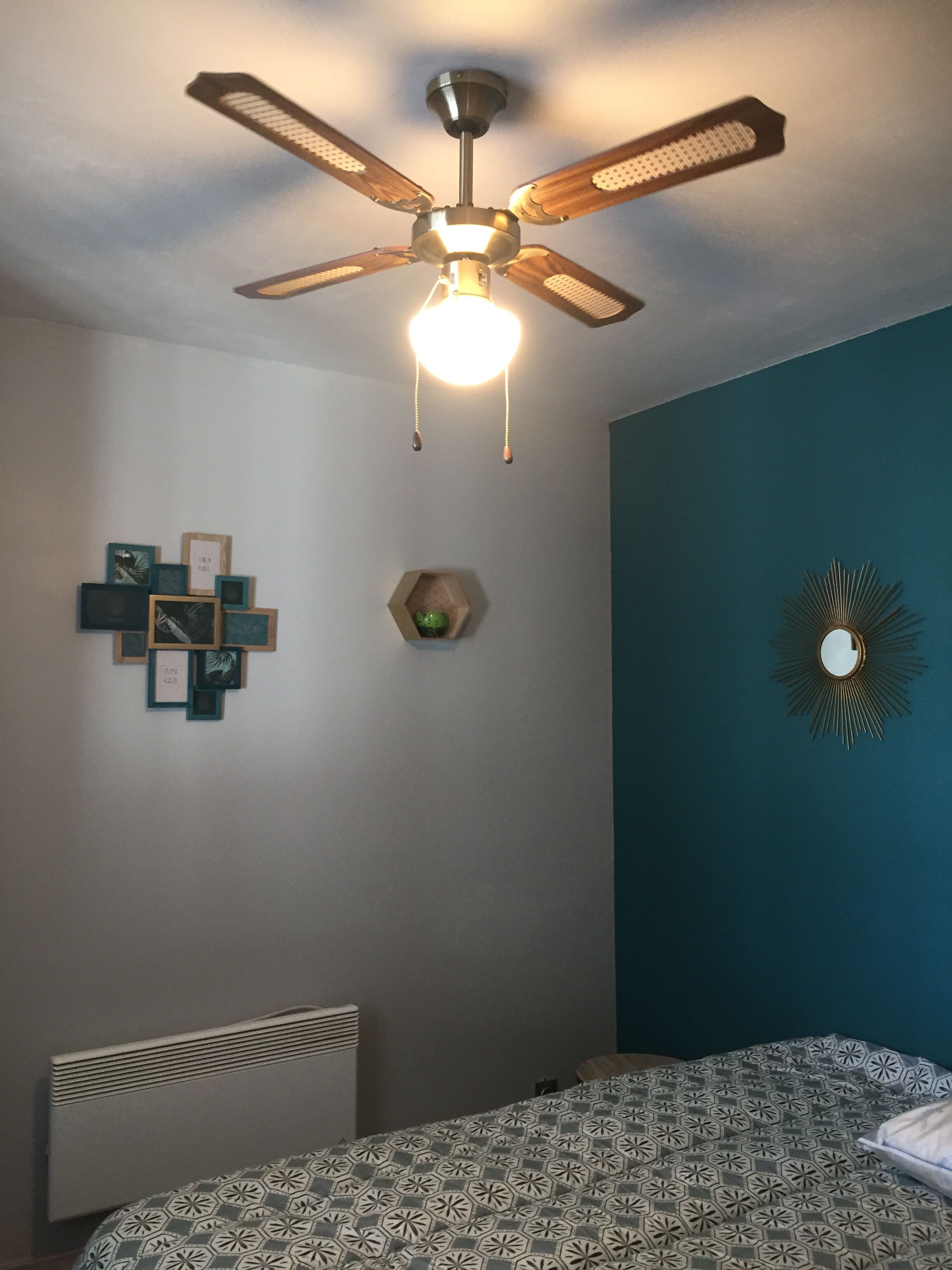 Een slaapkamer met een plafondventilator, een bed met beddengoed met een patroon, een radiator en wanddecoraties, waaronder een spiegel en fotolijstjes.