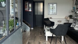 Un espace de vie moderne avec un plancher en bois, un coin repas avec une table blanche et des chaises noires, un canapé noir agrémenté de coussins à motifs et un mur de verre donnant sur l’extérieur.
